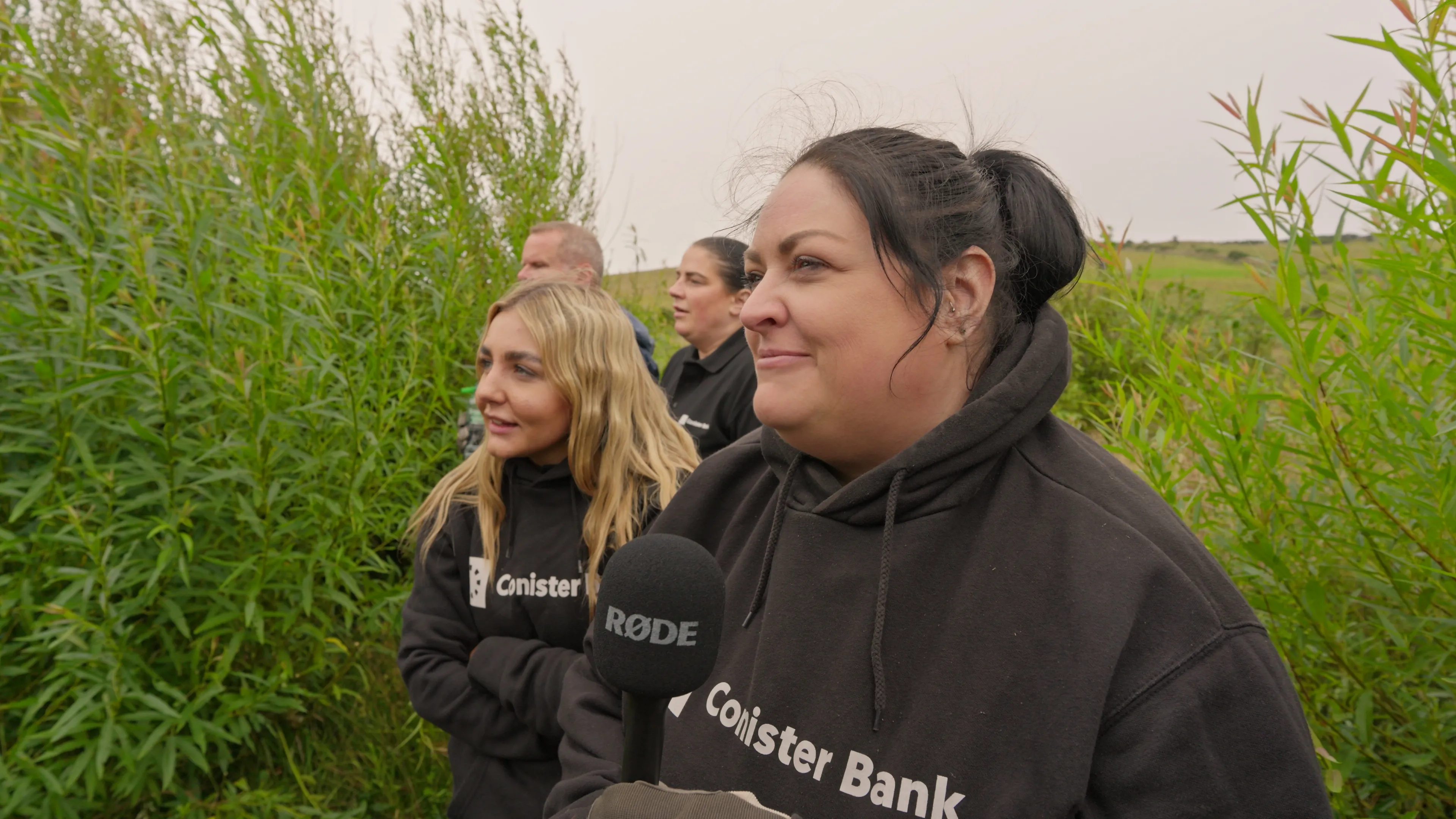 A group of people, wearing black Conister Bank hoodies, stand outdoors amid tall greenery. One person holds a microphone, smiling warmly. The mood is cheerful.
